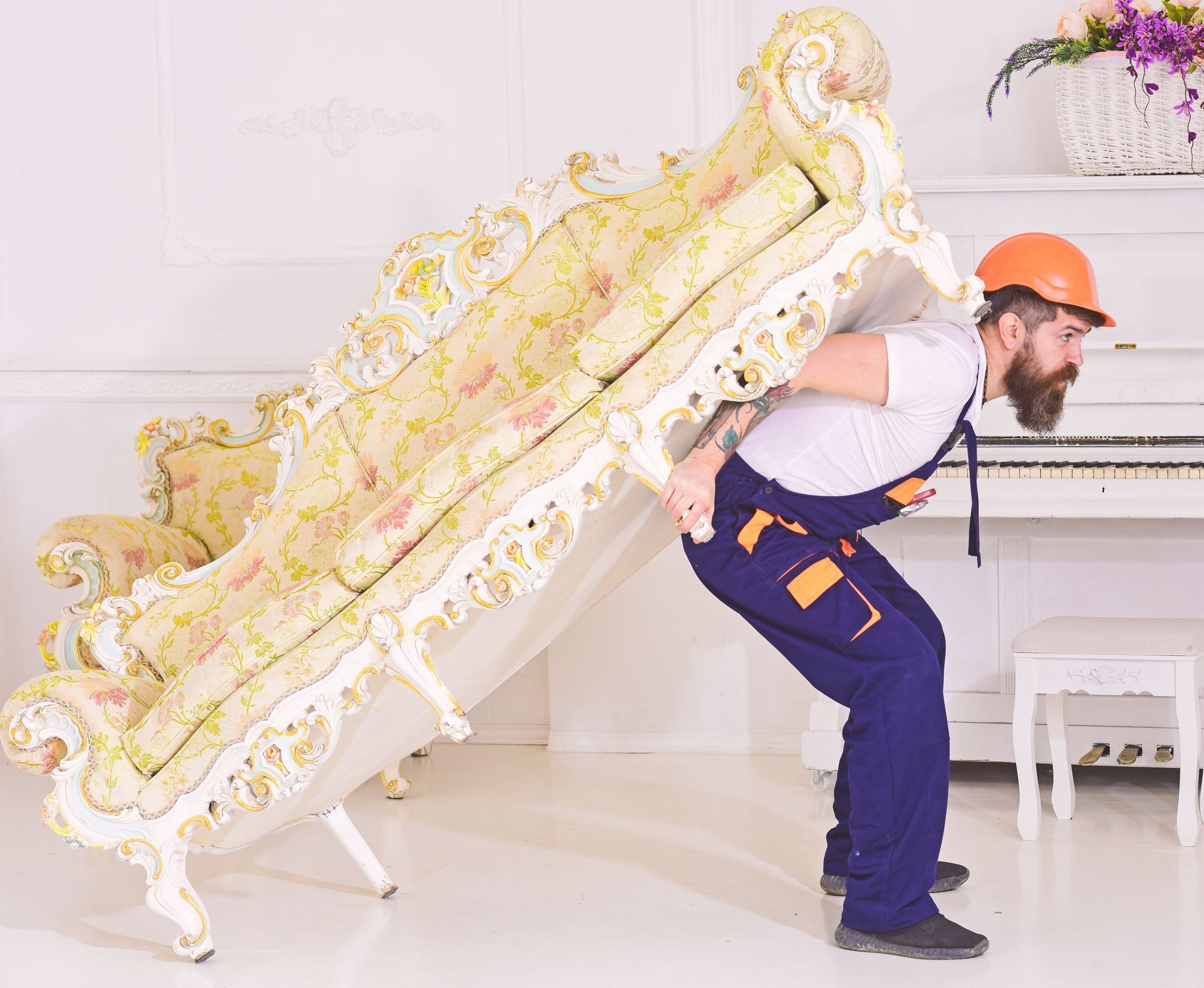 Relocation concept. Courier delivers furniture in case of move out, relocation. Loader moves sofa, couch. Man with beard, worker in overalls and helmet carries sofa on back, white background.